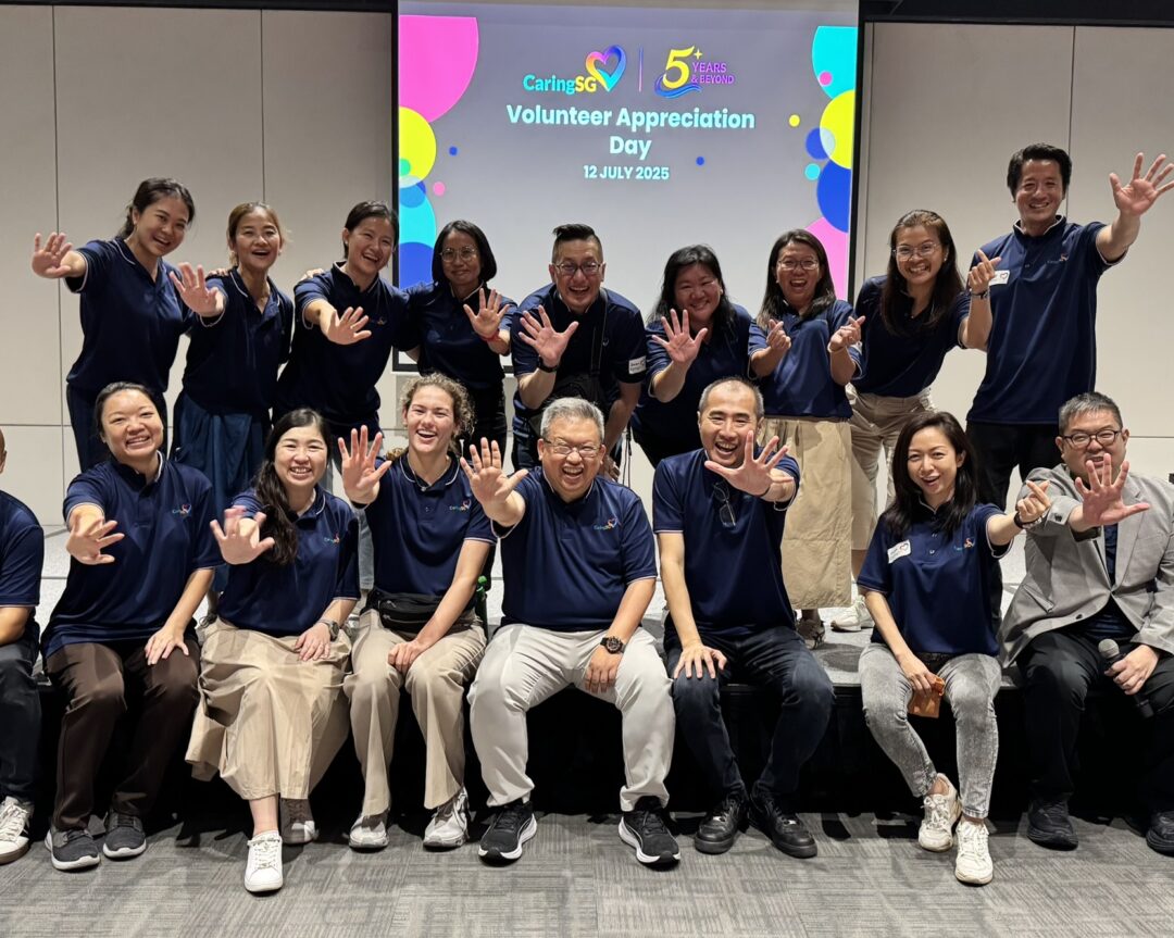 A group of CaringSG team members pose joyfully in two rows, all wearing matching navy blue shirts and holding up five fingers to celebrate CaringSG’s 5th anniversary. They are smiling warmly in front of a screen displaying “Volunteer Appreciation Day – 12 July 2025 – 5 Years & Beyond.” The atmosphere is cheerful and celebratory.