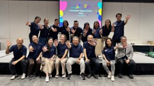 VAD2025 A group of CaringSG team members pose joyfully in two rows, all wearing matching navy blue shirts and holding up five fingers to celebrate CaringSG’s 5th anniversary. They are smiling warmly in front of a screen displaying “Volunteer Appreciation Day – 12 July 2025 – 5 Years & Beyond.” The atmosphere is cheerful and celebratory.