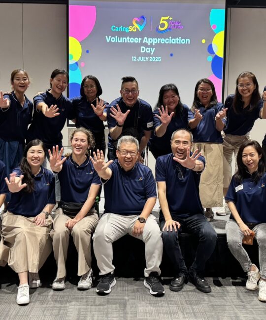 A group of CaringSG team members pose joyfully in two rows, all wearing matching navy blue shirts and holding up five fingers to celebrate CaringSG’s 5th anniversary. They are smiling warmly in front of a screen displaying “Volunteer Appreciation Day – 12 July 2025 – 5 Years & Beyond.” The atmosphere is cheerful and celebratory.